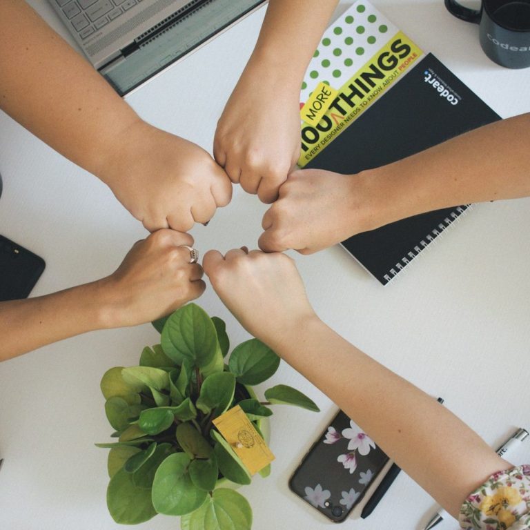 Fünf Hände treffen sich zur gemeinsamen Geste über Schreibtisch – Symbol für Teamzusammenhalt, Vertrauen und Zusammenarbeit
